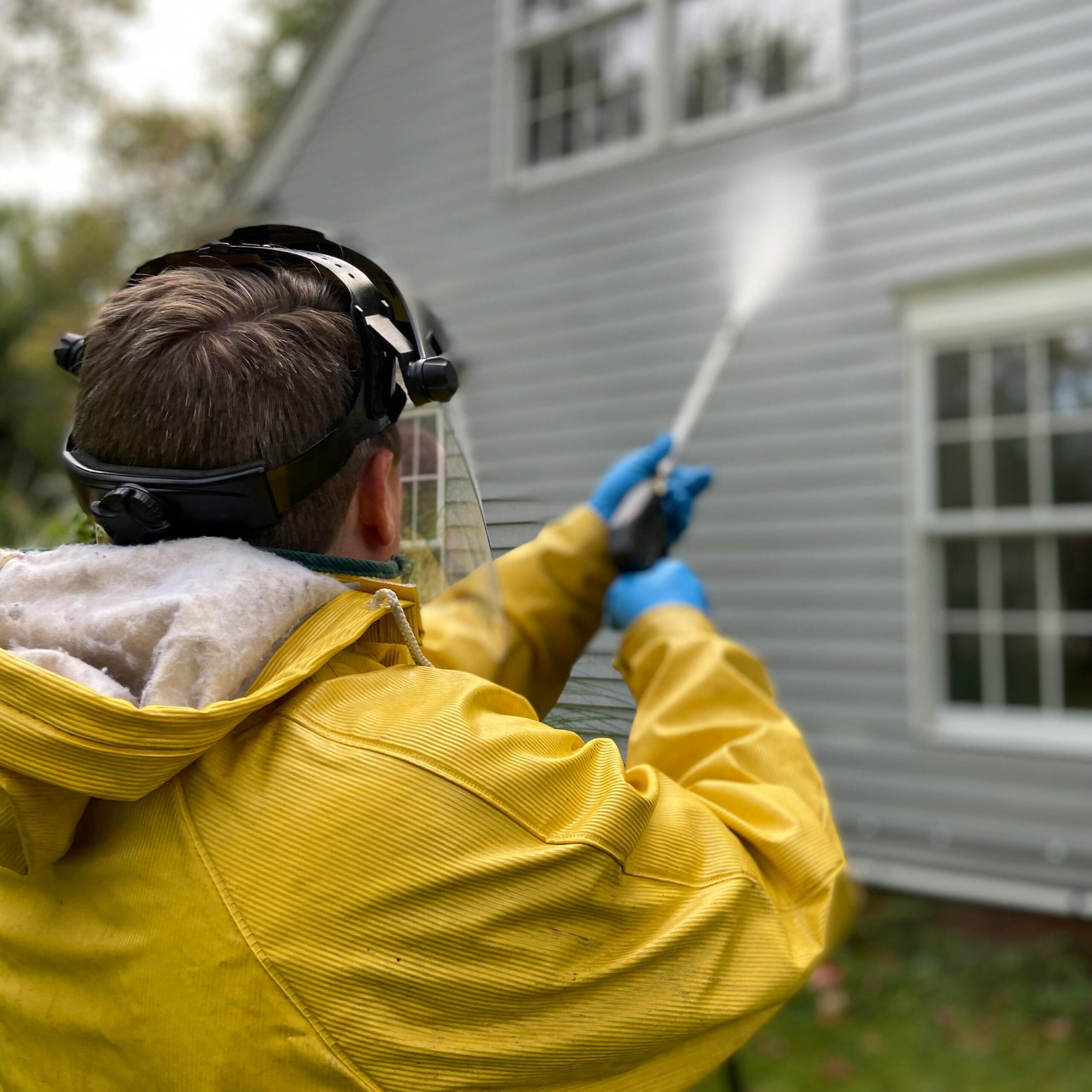 pexels photo 5652626 5652626 Person cleaning house siding with a pressure washer, wearing protective gear in a yellow raincoat.